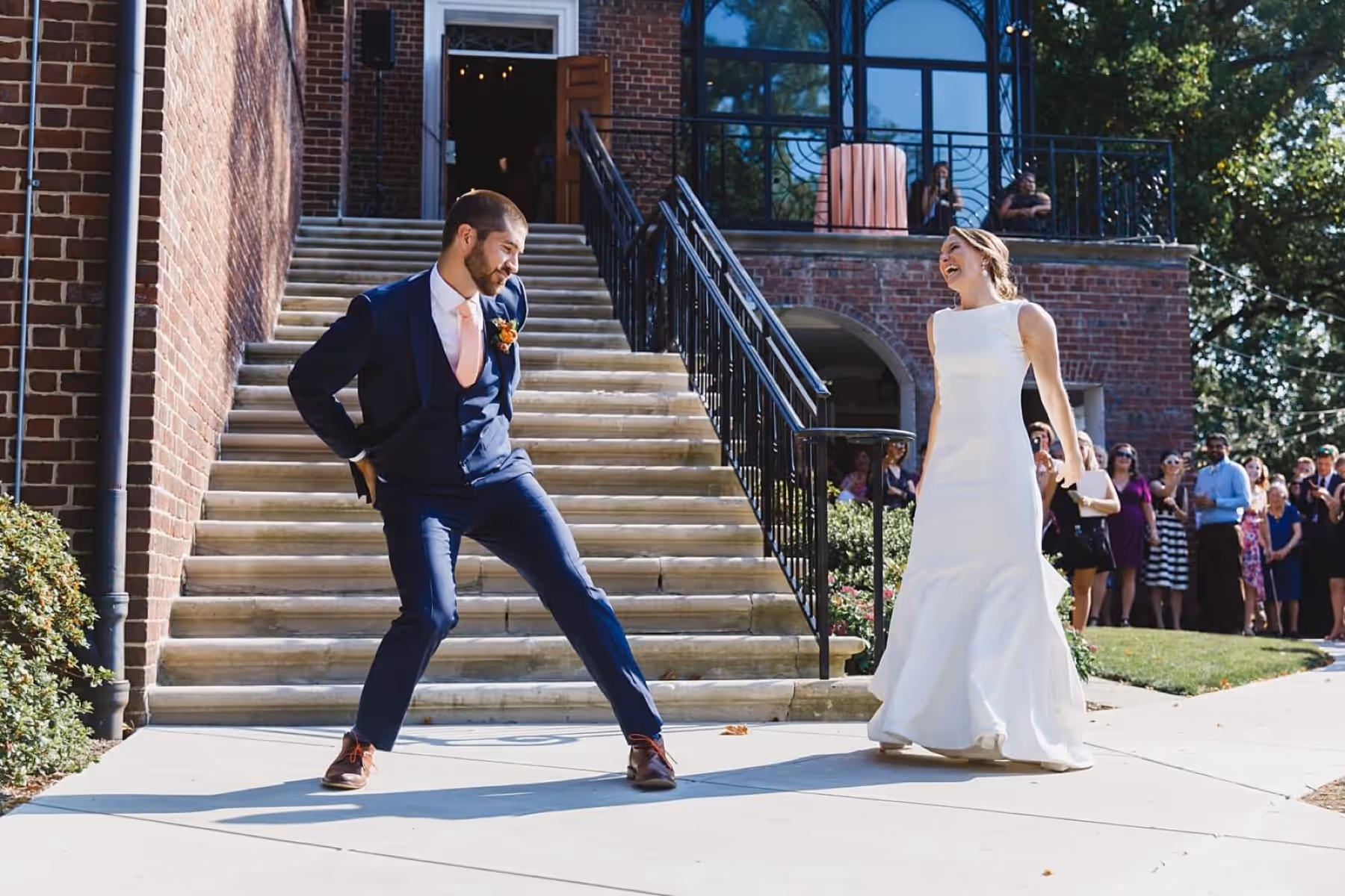 Couple dancing on the path outside their brick wedding venue while guests watch from the steps