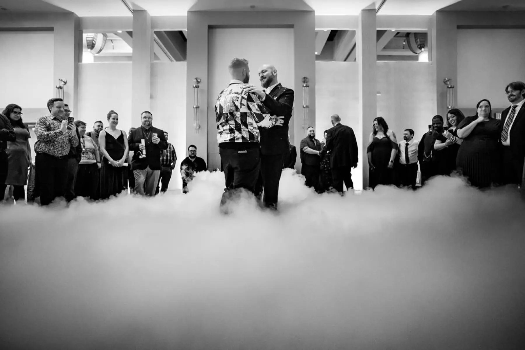 Two grooms dancing in low fog on a ballroom floor as guests watch, in black and white