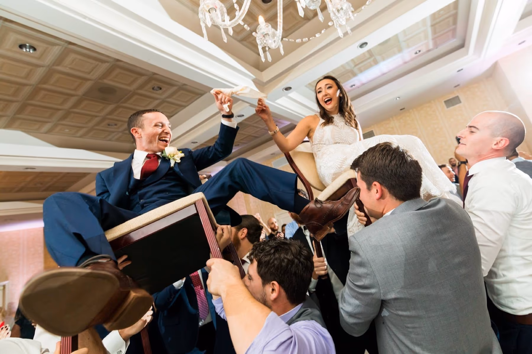 Groom and bride lifted on chairs by guests during a hora at a ballroom reception