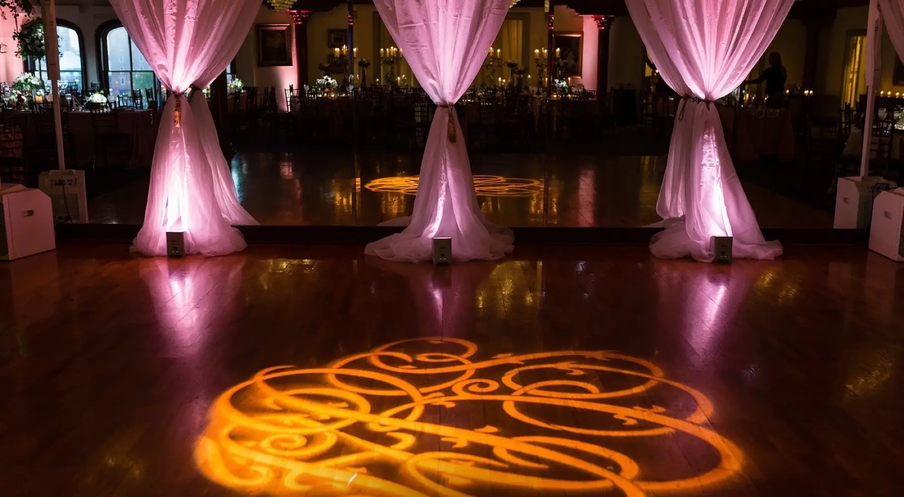 Gold monogram light projected on a wooden dance floor in front of pink-lit draped columns
