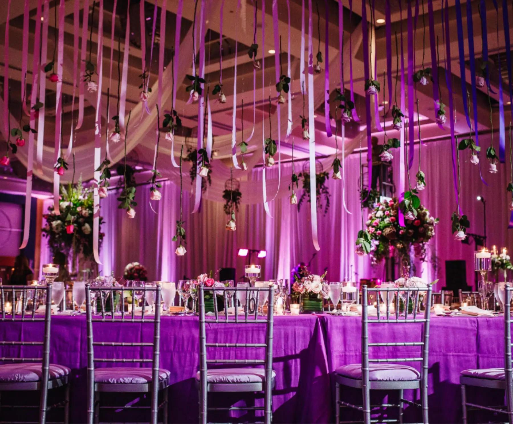 Ballroom washed in purple uplighting with hanging florals over the tables